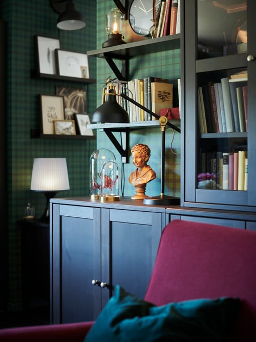 A black RANARP work lamp with an adjustable head and arm is on a cabinet, by an armchair and some shelves holding books.