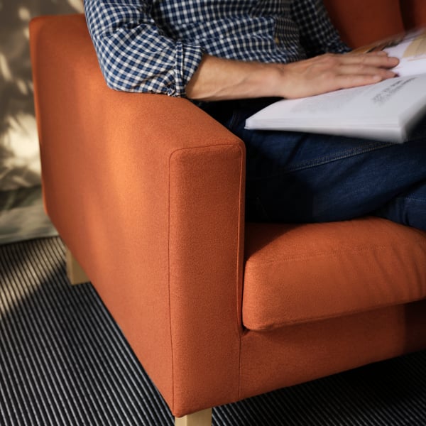 A red-brown SALTSJÖBADEN armchair with a person in a patterned shirt sitting comfortably in it, leafing through a magazine.