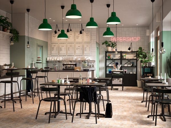 A café with black tables and chairs, green and white walls, a paper shop, green pendant lights and plants.