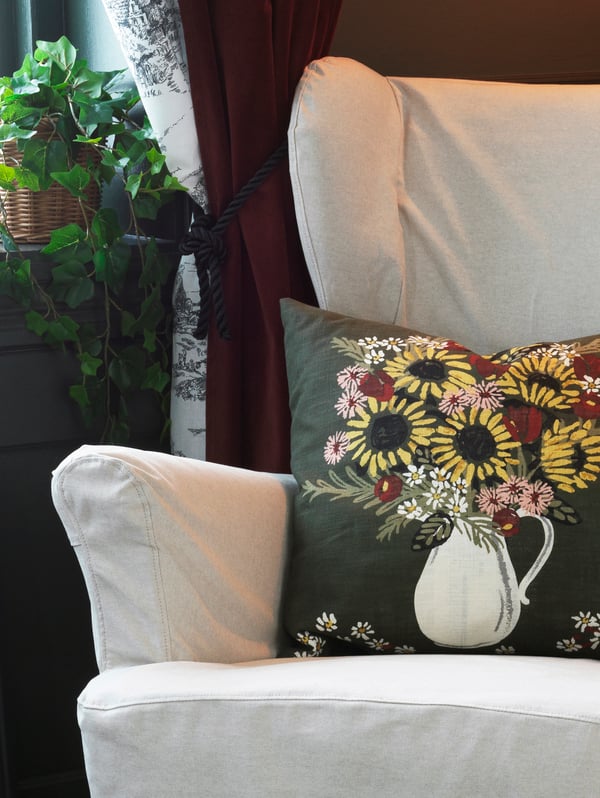 Poltrona em branco com almofada DEKORERA em verde e padrão floral. Uma planta junto a uma janela está atrás da cadeira.