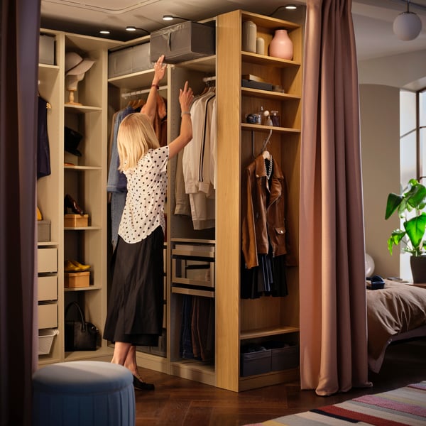 A person in a bedroom, reaching for a storage case in the upper section of an oak veneer PAX wardrobe.