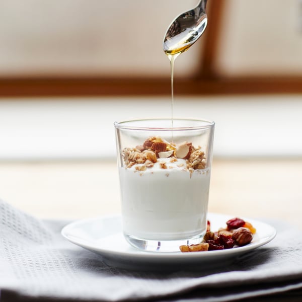 A spoon pours honey into an IKEA 365+ glass which contains yoghurt and muesli and stands on a white plate.