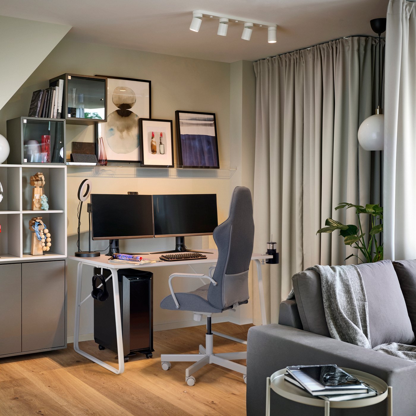 Vardagsrum med beige väggar och grå soffa. I bakgrunden syns ett vitt gamingbord och en grå gamingstol intill gönster med beige gardiner.