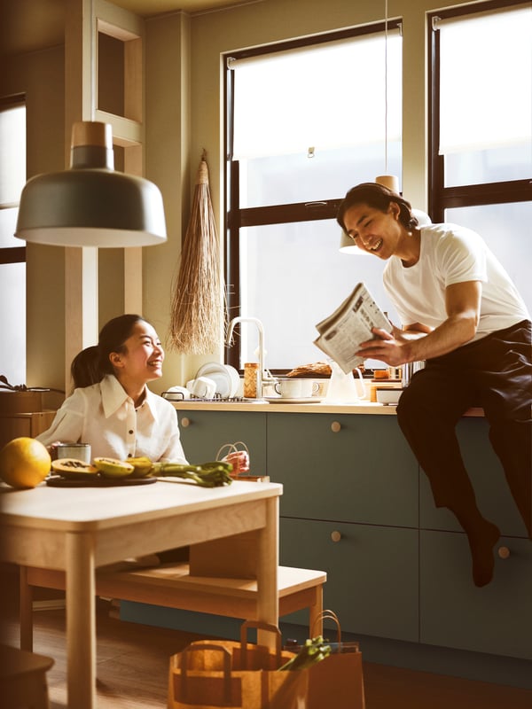 Two people chatting in a kitchen with grey-green BODARP drawer fronts, cabinets, a worktop, a sink and a table with a bench.