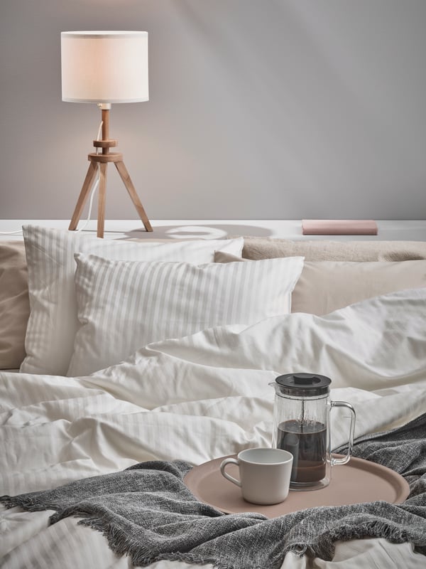 Bedroom area with bed covered in NATTJASMIN duvet covers and pillows with an INGRUN throw with a food tray on top of the bed with a IKEA 365+ mug and EGENTLIG jug on top. A LAUTERS table lamp stands at the bedhead letting off a low glow. 