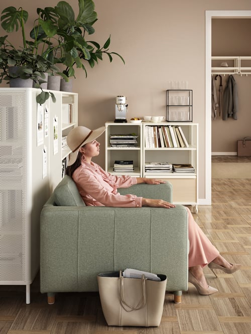 A woman sits in a Gunnared light green/wood LANDSKRONA armchair in an office waiting area, by shelves with books and drinks.