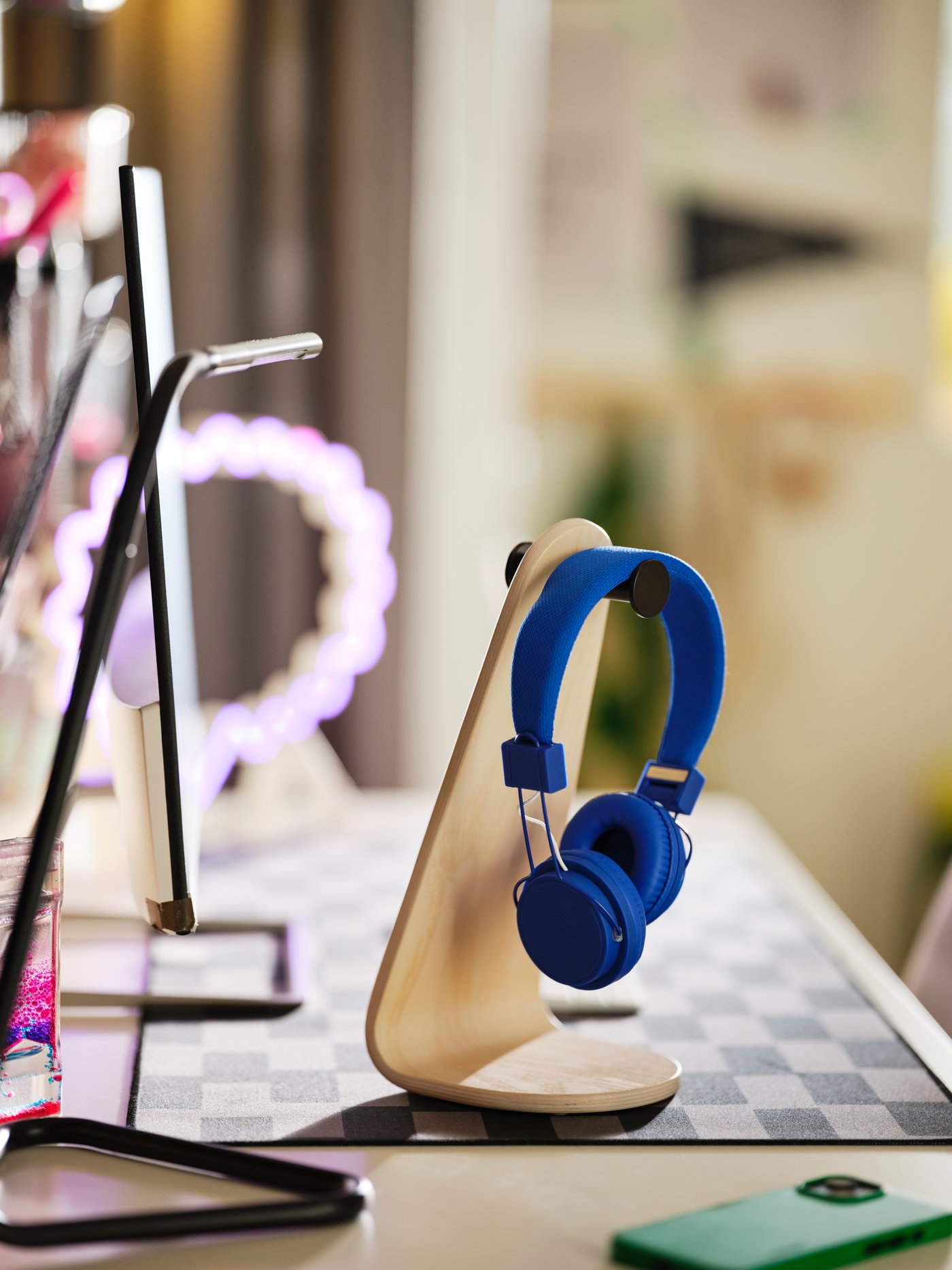 Sobre un escritorio, un soporte de auriculares LÅNESPELARE de chapa de fresno sostiene un par de auriculares de color azul brillante en su percha de metal negro.