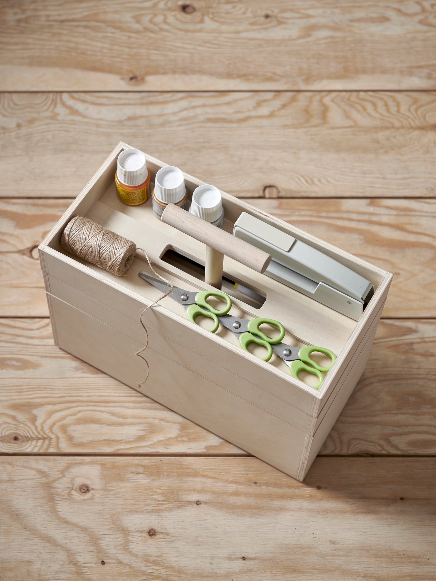 A wooden storage box with a handle and three levels holding scissors, string, paint and a stapler on the top level.