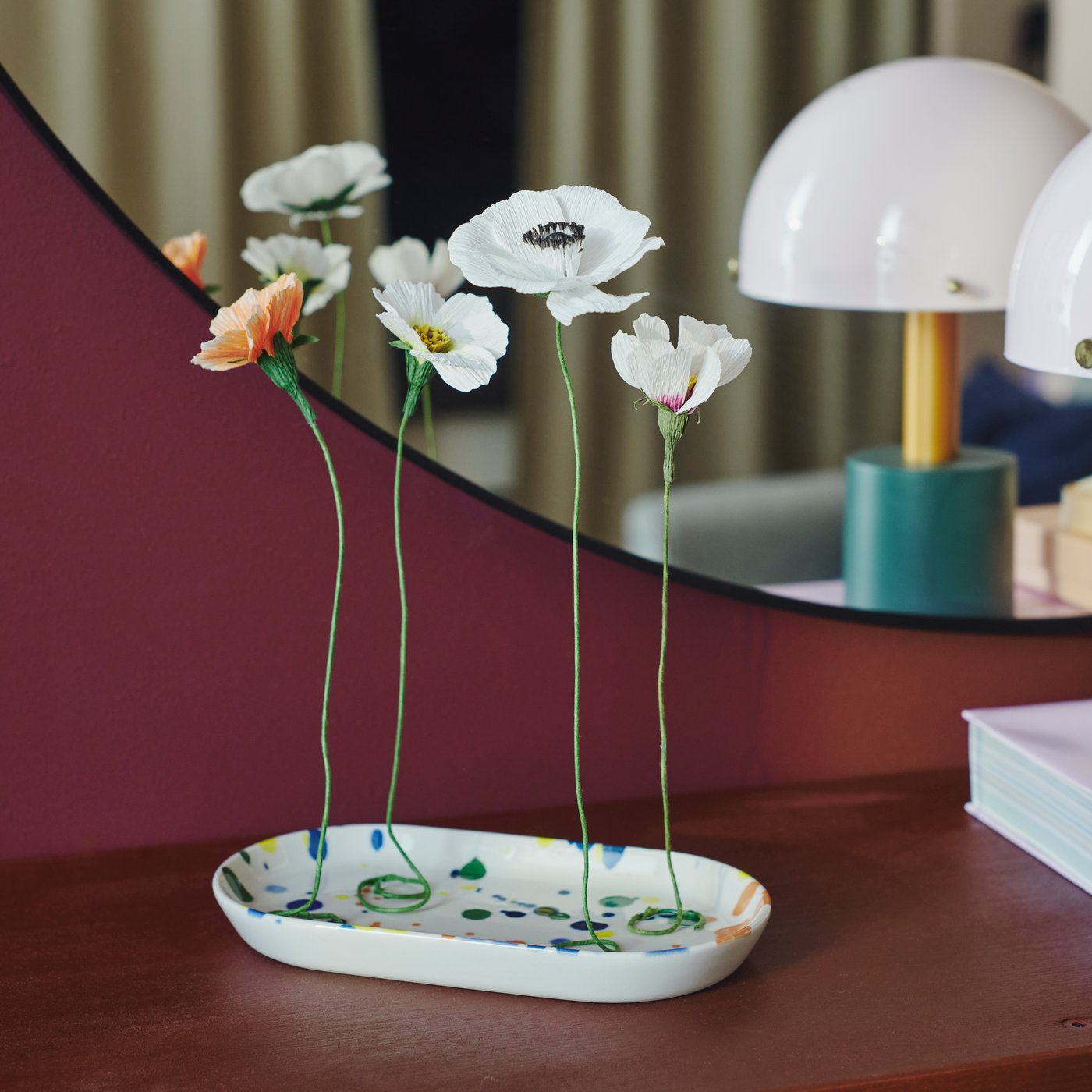 Några GULDSPETT pappersblommor står på ett färgglatt RÖDSNULTRA serveringsfat framför en LINDBYN spegel.