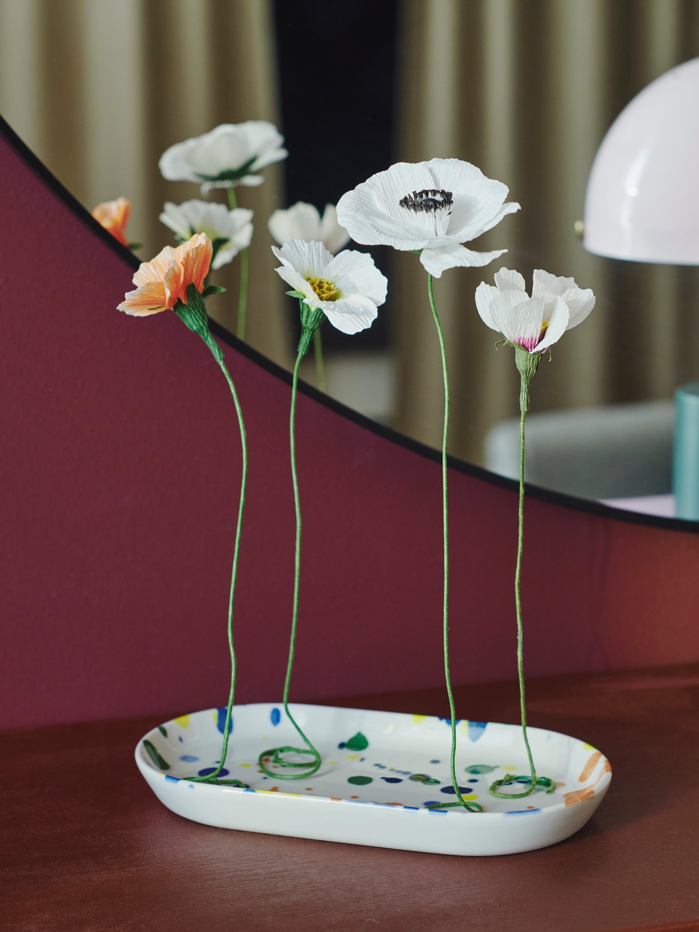 GULDSPETT Kunstblumen arrangiert auf einer bunt gestalteten RÖDSNULTRA Servierplatte, platziert vor einem LINDBYN Spiegel.