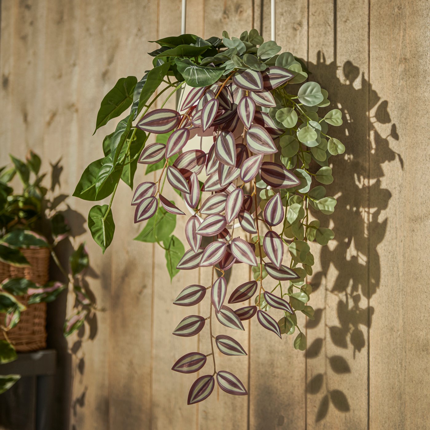 Un macetero colgante CHILISTRÅN con plantas artificiales en maceta FEJKA dentro, cuelga en un balcón soleado con paredes de madera.