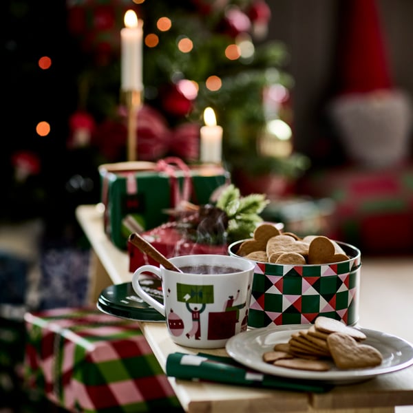 VINTERSAGA heart-shaped ginger thins are in a tin container and a plate next to a VINTERFINT mug, on a bench with presents.