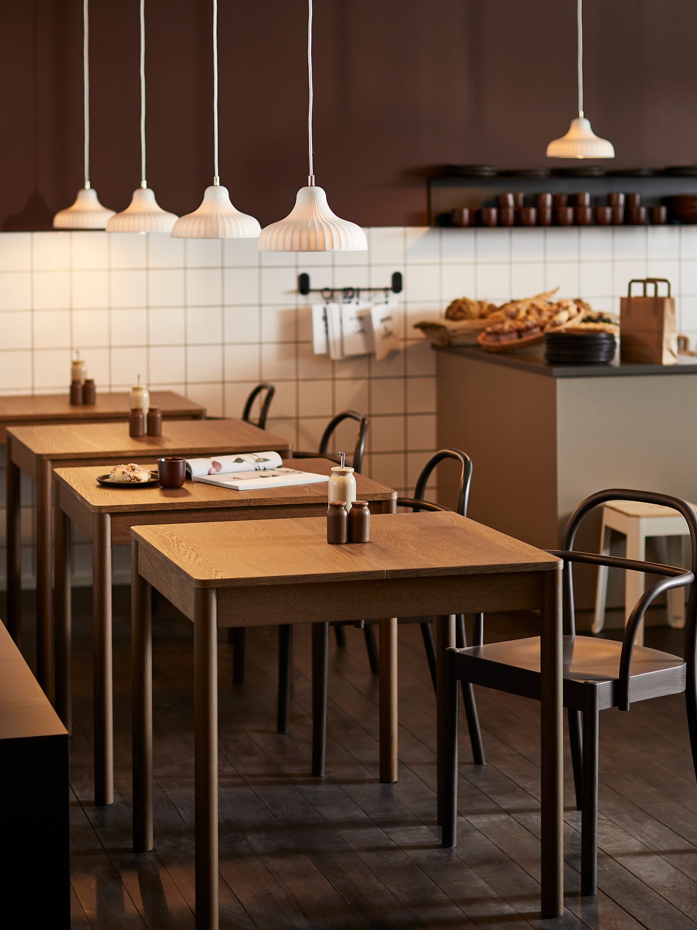 Sala di un ristorante dal tono caldo con tavoli in legno chiaro allineati, sedie nere, lampade a sospensione bianche sopra e bancone di servizio sullo sfondo con pane e utensili da cucina.