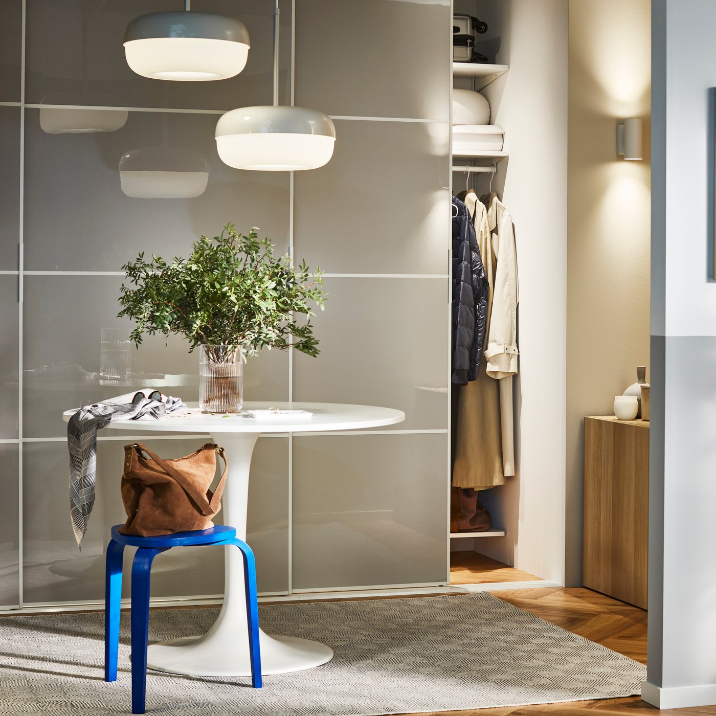Un tavolo rotondo e uno sgabello KYRRE blu in un ingresso, davanti a una combinazione di guardaroba SKYTTA/HOKKSUND con ante scorrevoli.