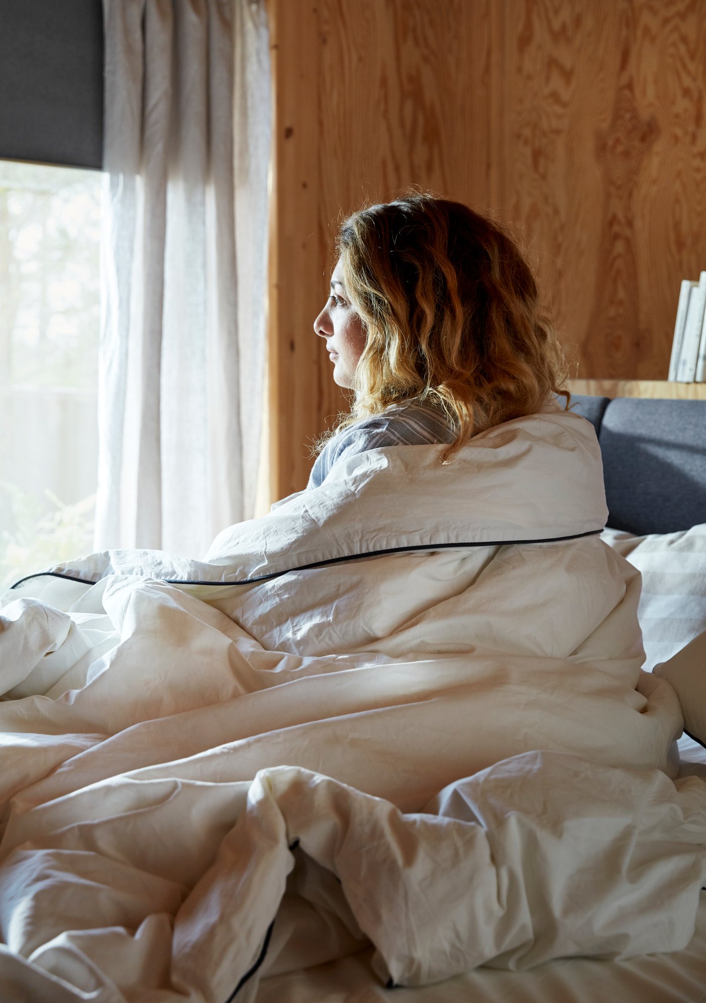 Nainen katselee ikkunasta ulos kietoutuneena muhkean peiton sisälle.