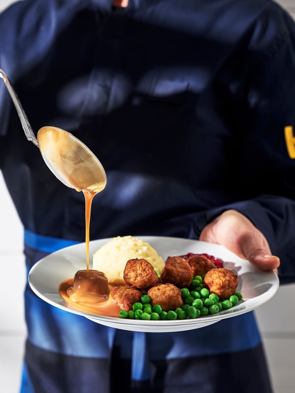 En person iført et blåt IKEA forklæde øser med en suppeske kartoffelmos, grønne ærter og creme sauce samt en tyttebærsauce og 8 kyllingeboller op på en hvid tallerken.
En person iført et blåt IKEA forklæde øser med en suppeske kartoffelmos, grønne ærter og creme sauce samt en tyttebærsauce og 8 kyllingeboller op på en hvid tallerken.