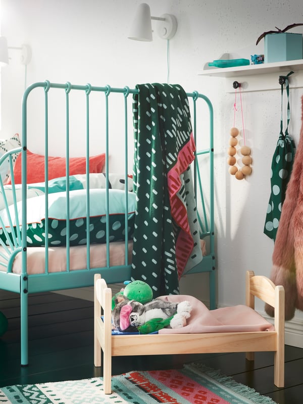 A solid pine DUKTIG doll’s bed with bed linen and soft toys stands under a shelf and next to a MINNEN extendable bed.