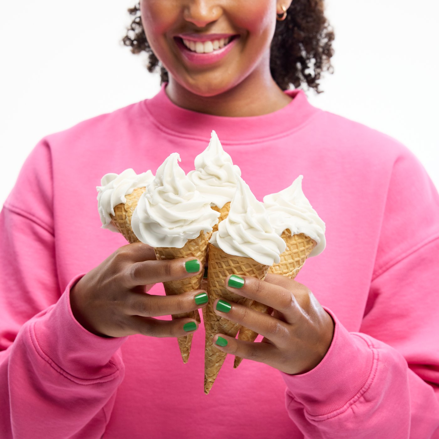 A smiling woman in a pink sweater and green nail polish, happily struggles to carry five soft ice cream cones in her hands.
