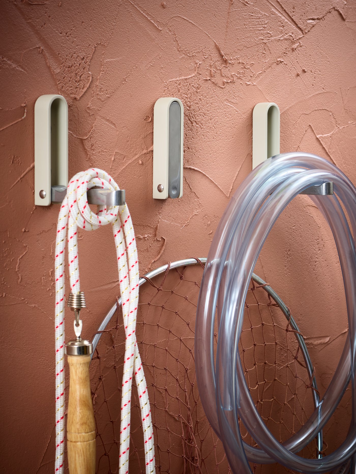 Three beige KLYKET folding hooks with a jump rope, a fishing net and a hose on a gritty, dusted pink wall.