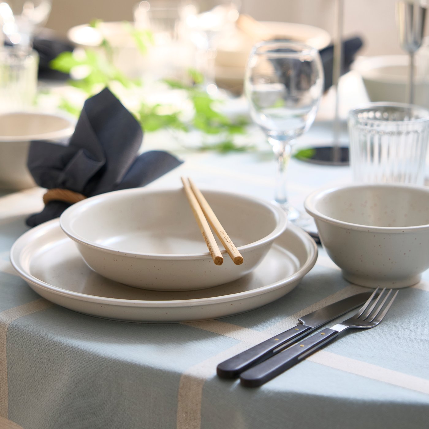 A dotted/light beige DAGGASTER bowl is placed on a plate, with two chopsticks on top of the bowl and cutlery on the side.