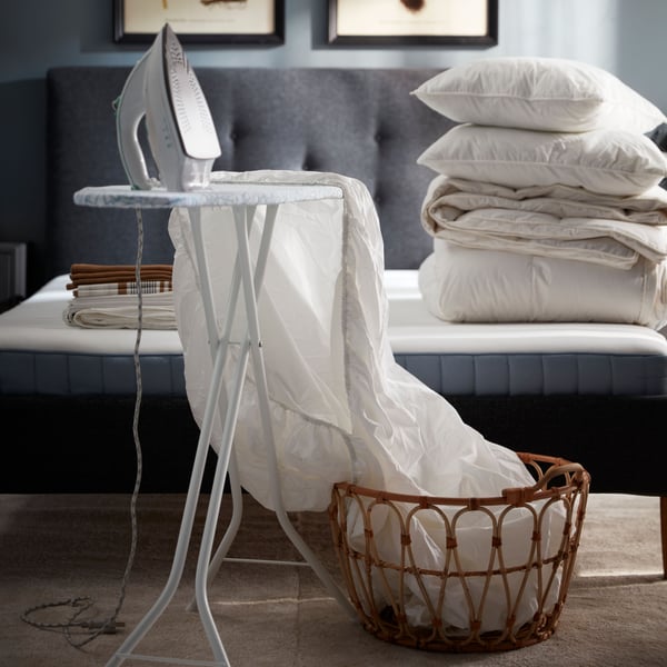 A white NATTJASMIN fitted sheet that is half in a baske and half laid out on a RUTER ironing board in front of an unmade bed.