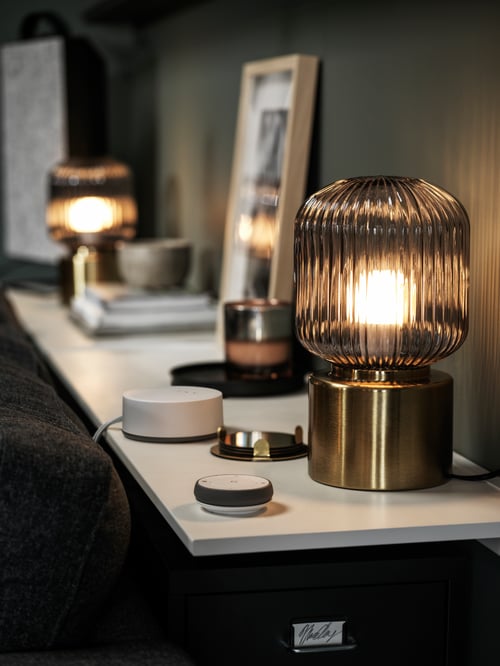 Une étagère murale derrière un canapé avec des lampes de table en laiton SOLKLINT, un chargeur LIVBOJ, une télécommande TRÅDFRI et une décoration.