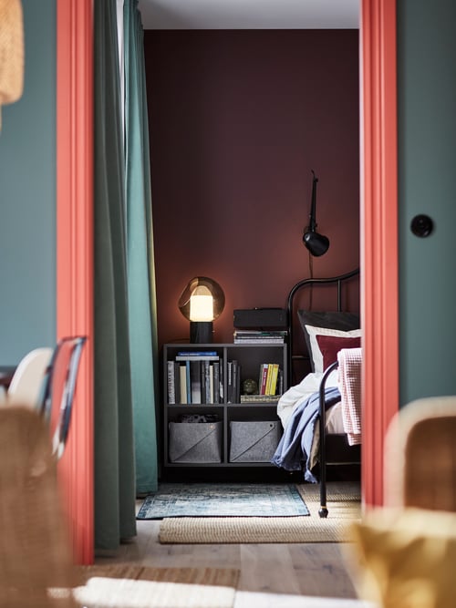 Bedroom with a table lamp resting on a cabinet that contains multiple books and two baskets, with a bed on the right.
