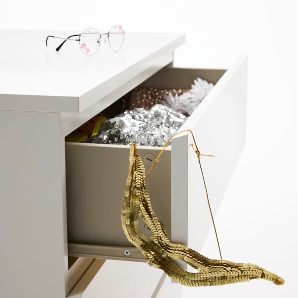 Close-up of a white MALM chest of 4 drawers. A drawer is open, showing a silver sequin dress and a gold masquerade mask.