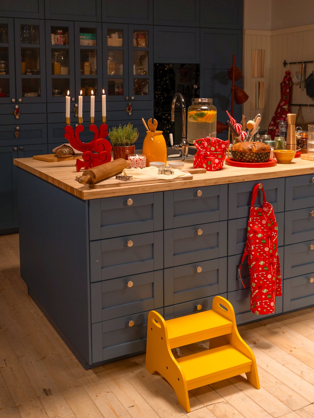 Un'isola da cucina blu con un piano di lavoro in legno, preparata per i dolci di Natale. Sul piano ci sono un panettone, un candelabro rosso VINTERFINT e uno sgabello giallo BEKVÄM in primo piano.