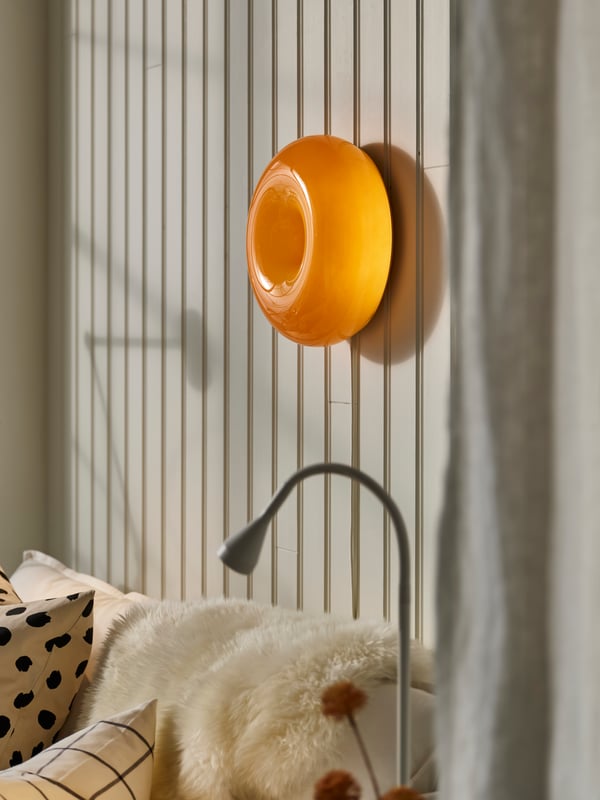 A round, orange VARMBLIXT wall lamp over a white sofa with an off-white ULLERSLEV sheepskin, plus a white NÄVLINGE work lamp.