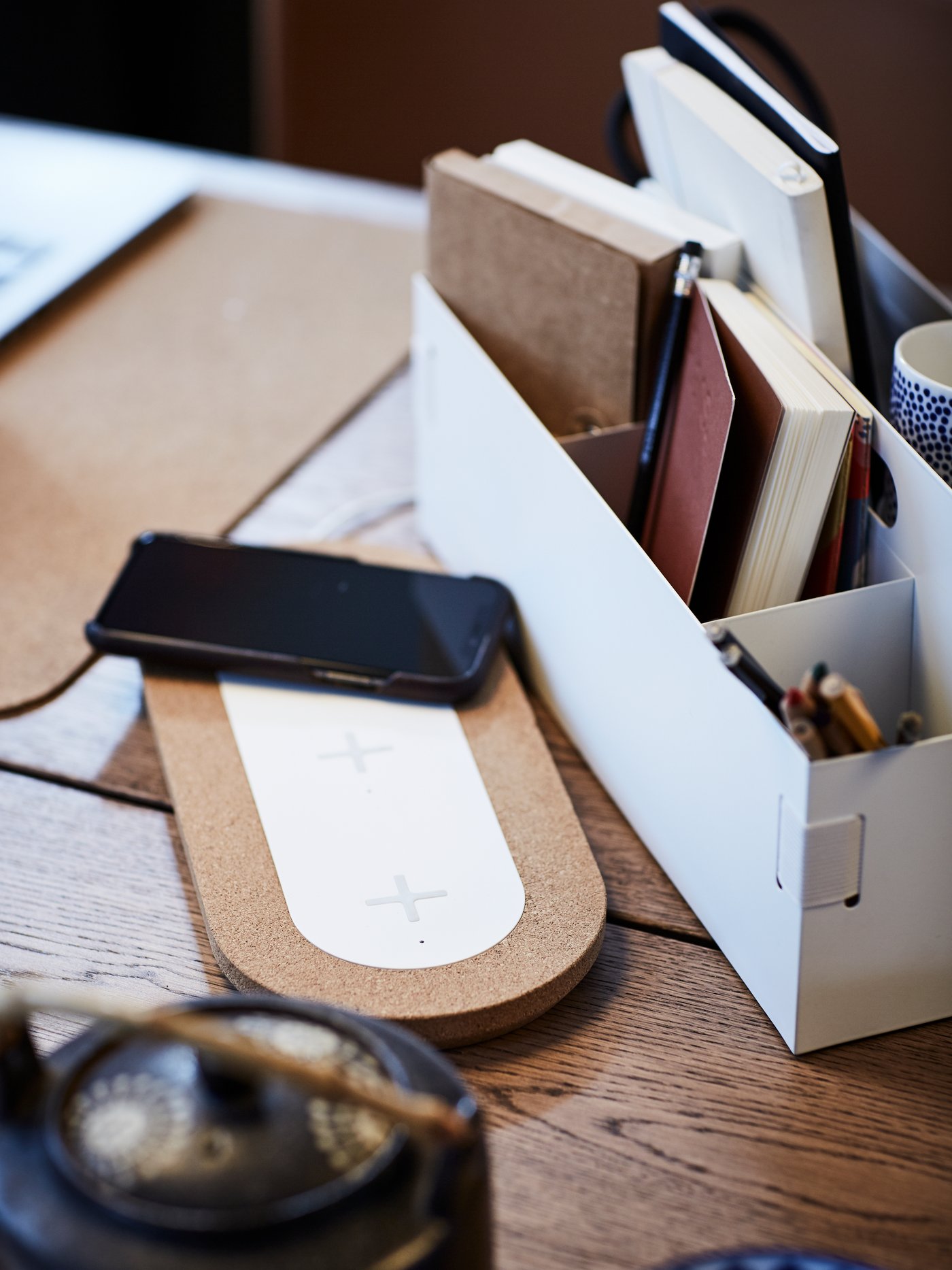 Notebooks in a KVISSLE desk organiser, next to a mobile phone on top of a NORDMÄRKE triple pad for wireless charging.