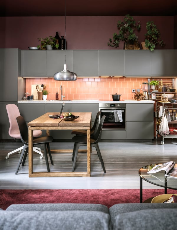 Eine dunkelgraue Küche mit rosa Fliesenspiegel, anthrazitfarbenen Stühlen, einem Esstisch aus Eichenfurnier und einer VÄXJÖ Hängeleuchte.