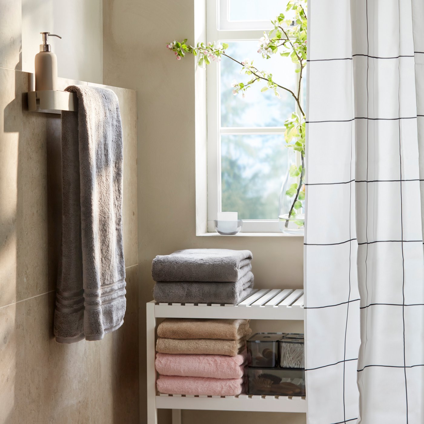 A bathroom corner has grey BROKGLIM hand towels stacked on a white bench by a window, a towel rail and a shower curtain.