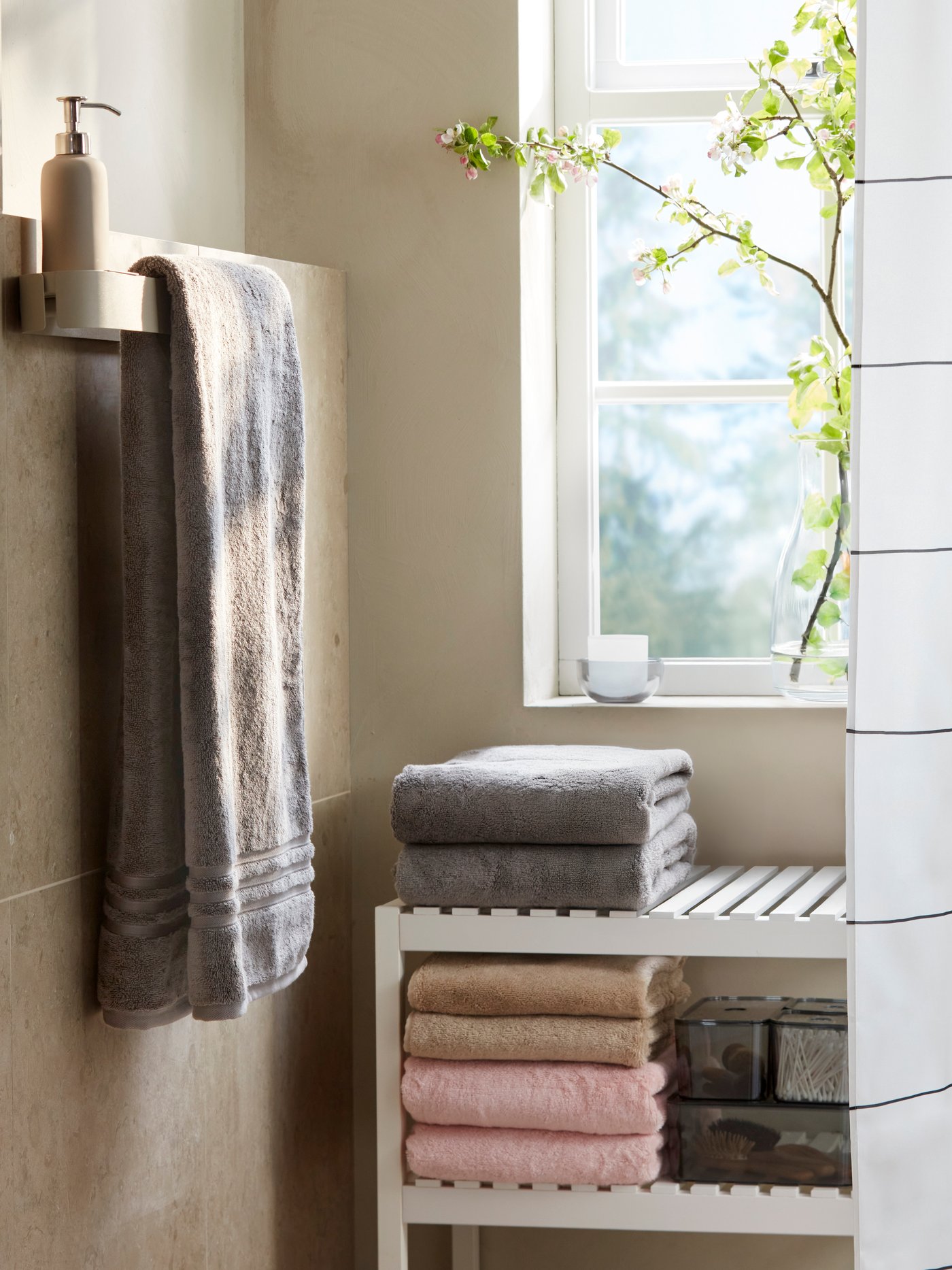 A bathroom corner has grey BROKGLIM hand towels stacked on a white bench by a window, a towel rail and a shower curtain.