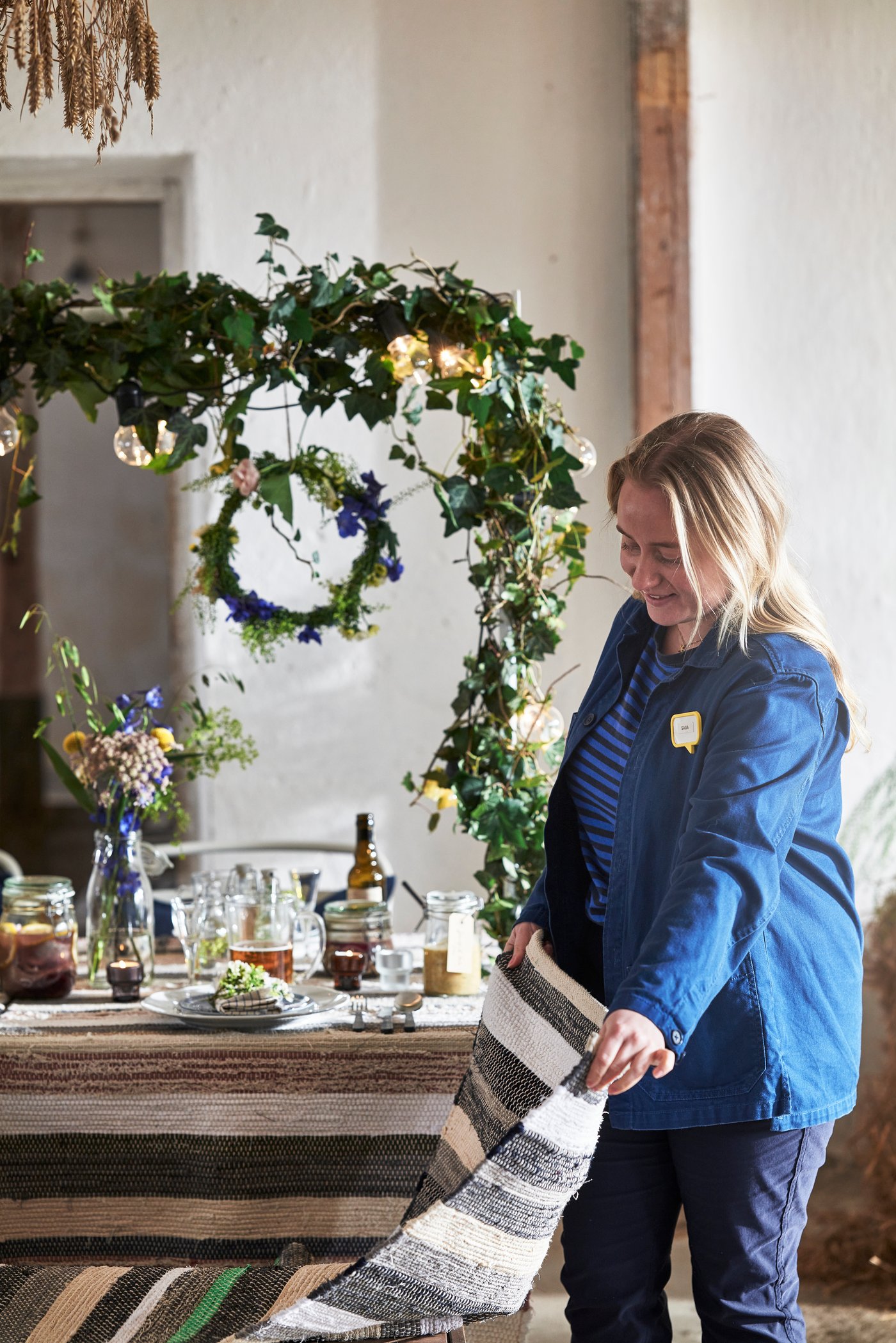 En person står vid ett rustikt dukat bord och arrangerar en randig textil; bordet är dekorerat med blommor, gröna blad och tända ljus, med en spegel och grönskande kransar i bakgrunden.
