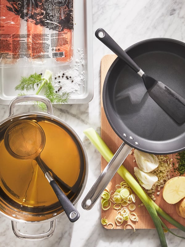 An empty IKEA 365+ frying pan with a black turner is placed on a chopping board and there is a pot with a ladle next to it.