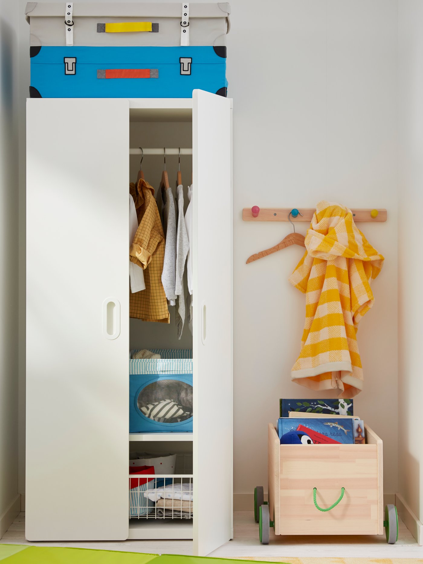 ארון בגדים STUVA/FRITIDS פתוח למחצה ניצב לצד מתלה עם ידיות FLISAT המורכב על קיר וארגז אחסון עם גלגלים FLISAT.