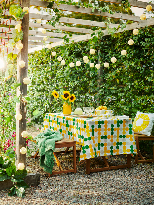 Ambiente esterno con tavolo apparecchiato con tovaglia BRÖGGAN. Sul tavolo ci sono girasoli e piante verdi ovunque.