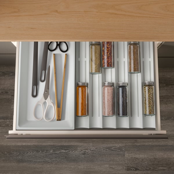 Offene Küchenschublade mit weißen UPPDATERA Einsätzen: Links liegen Schere, Kochzange und Essstäbchen, rechts stehen in Reihen mehrere CITRONHAJ Glasgewürzbehälter ordentlich auf einem gerillten Einsatz, unter einer Arbeitsplatte in Holzoptik.