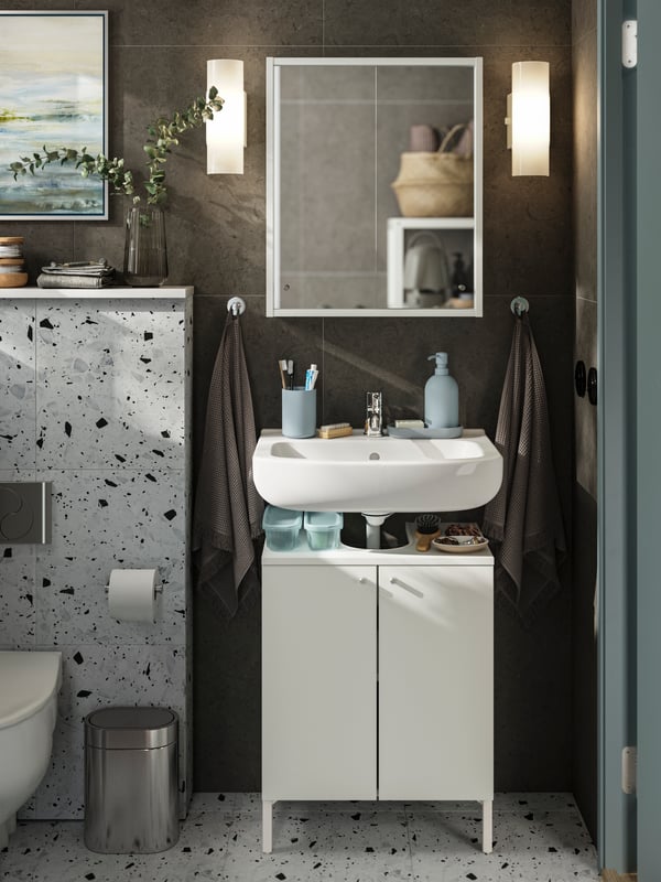 A NYSJÖN cabinet with two doors stands under a white sink with light grey-blue accessories on top and a mirror on the wall.