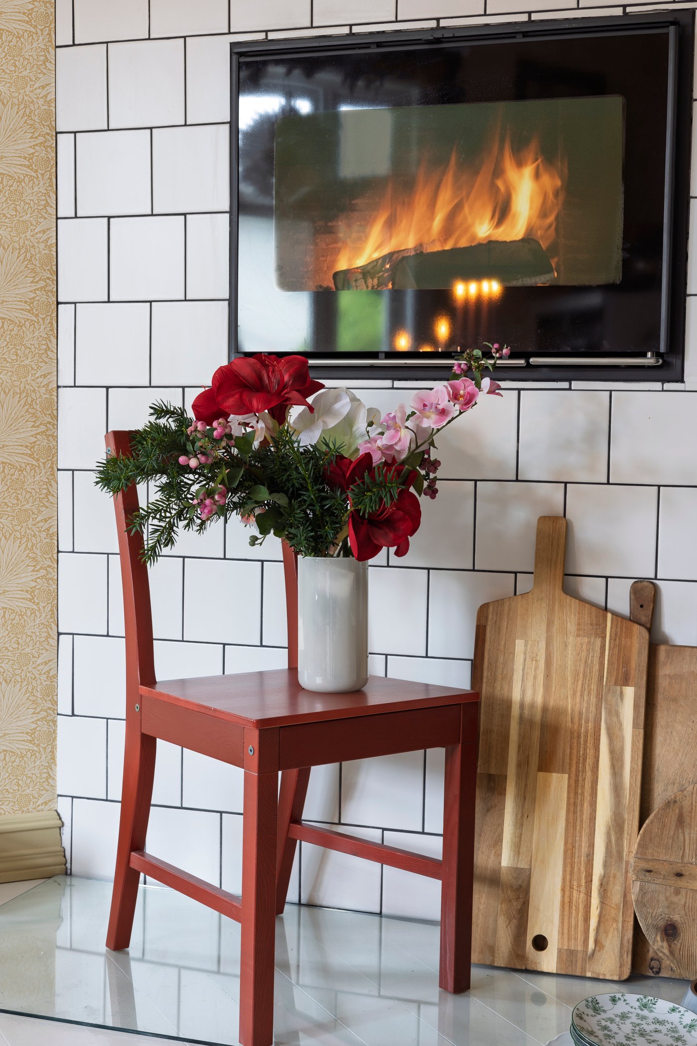 Röd stol med blomsterarrangemang framför kaklad vägg och brasa i bakgrunden.