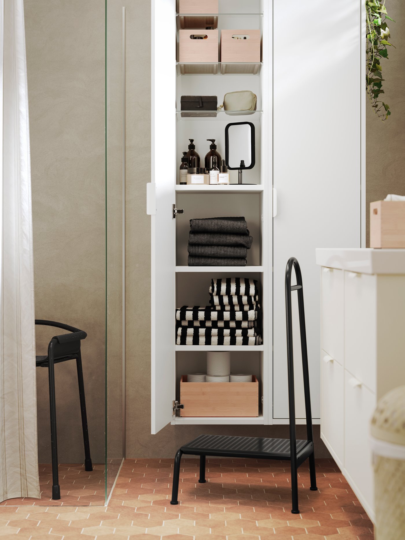 A BÄSINGEN step stool with handrail in anthracite is placed in a brown bathroom in front of a white HAVBÄCK high cabinet.