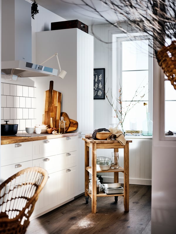 Dapur gaya desa berwarna putih dengan permukaan kerja kayu dan kabinet berwarna putih, dan troli BEKVÄM di tengah ruang.