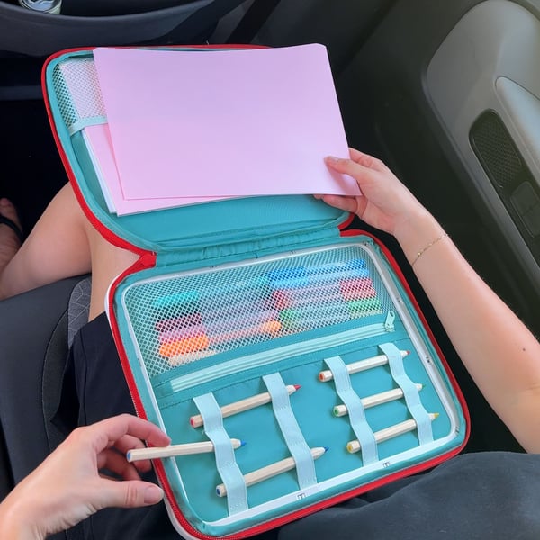 A child is sitting in a car and holding a MÅLA drawing case with MÅLA accessories.