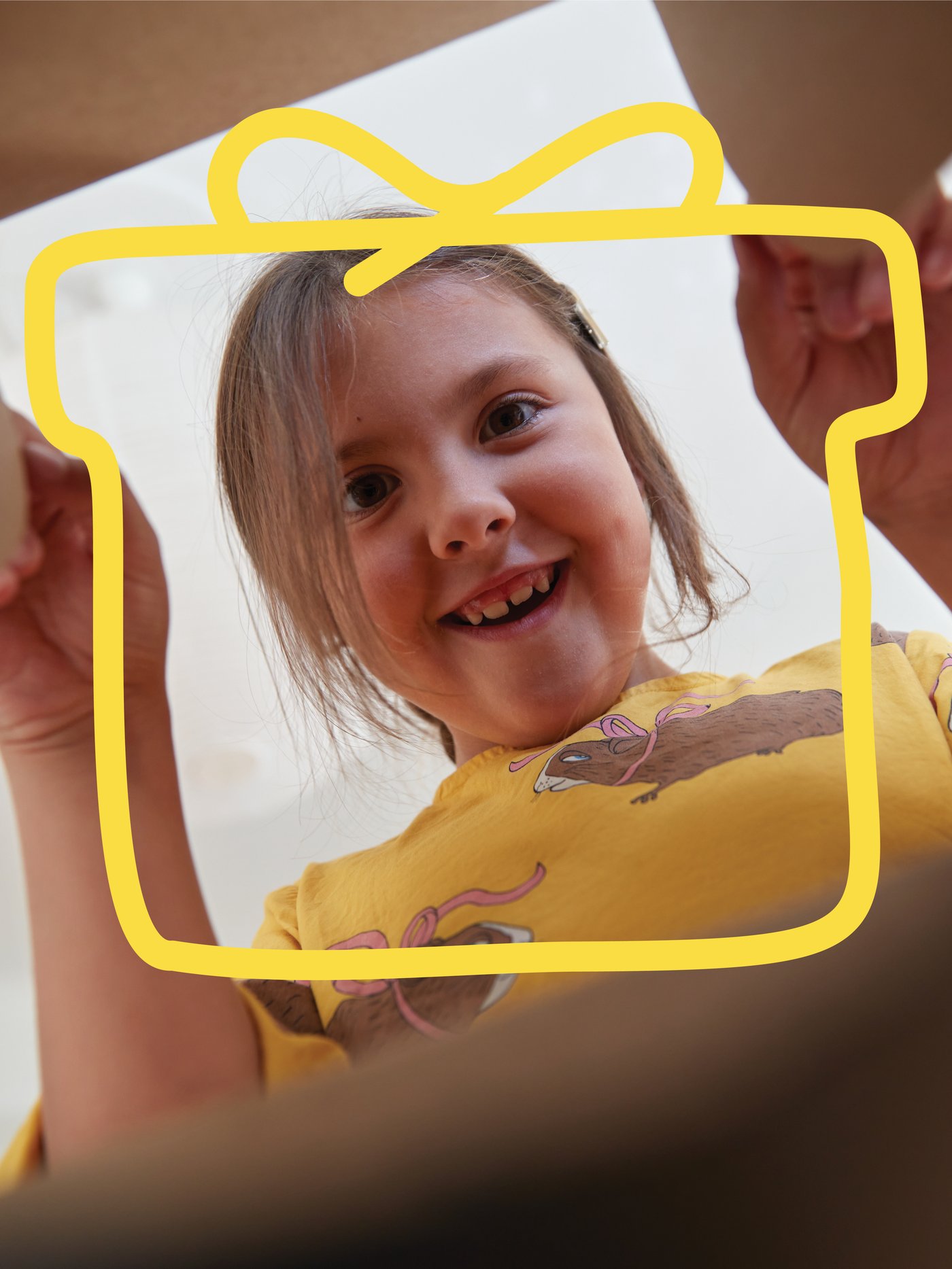 A little girl looking into a box. A yellow frame in the shape of a present on top of the image.