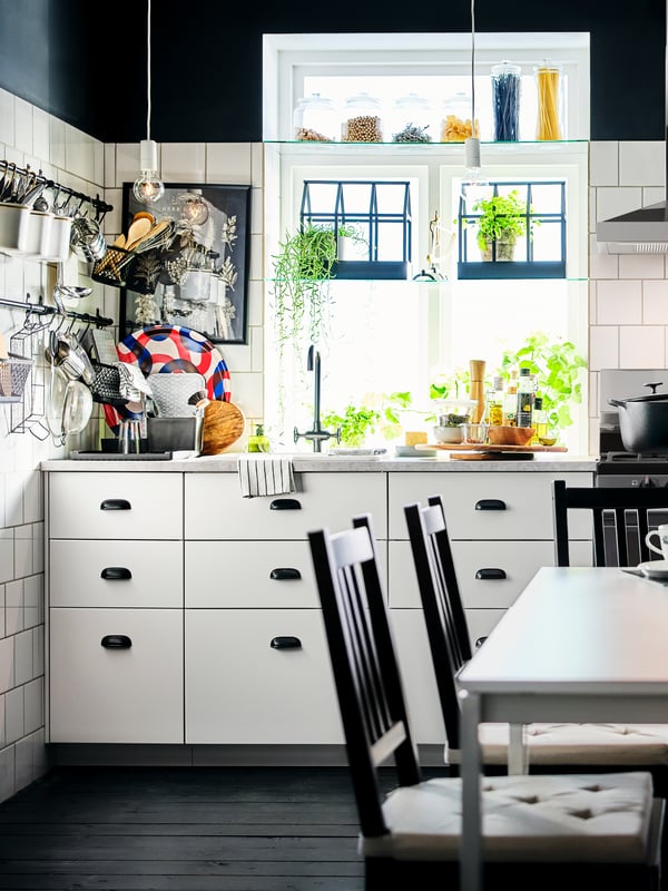 A corner of a black-and-white kitchen with white base cabinets, plants in the window and utensils on wall-mounted rails.