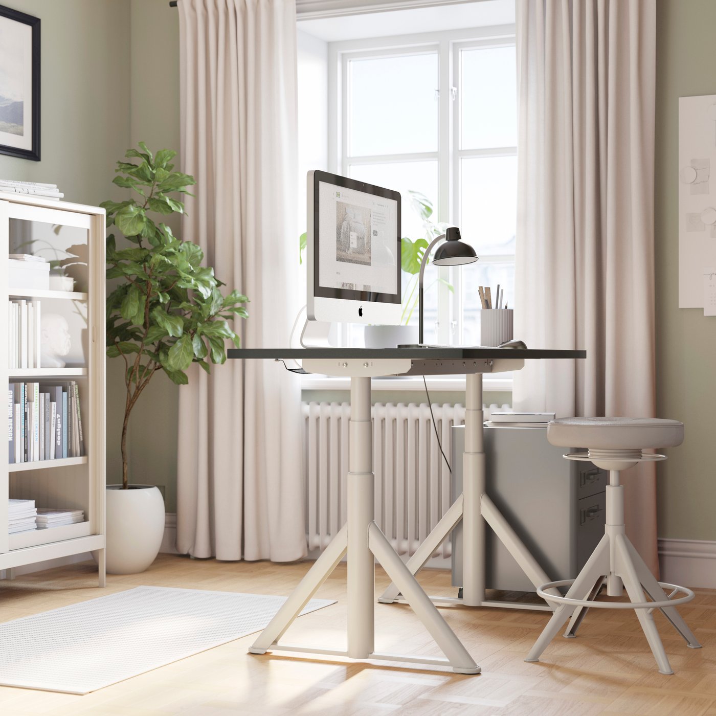 In un luminoso ufficio c'è una scrivania IDÅSEN color beige, pensata per lavorare sia in piedi che seduti, con accanto uno sgabello rotondo IDÅSEN.