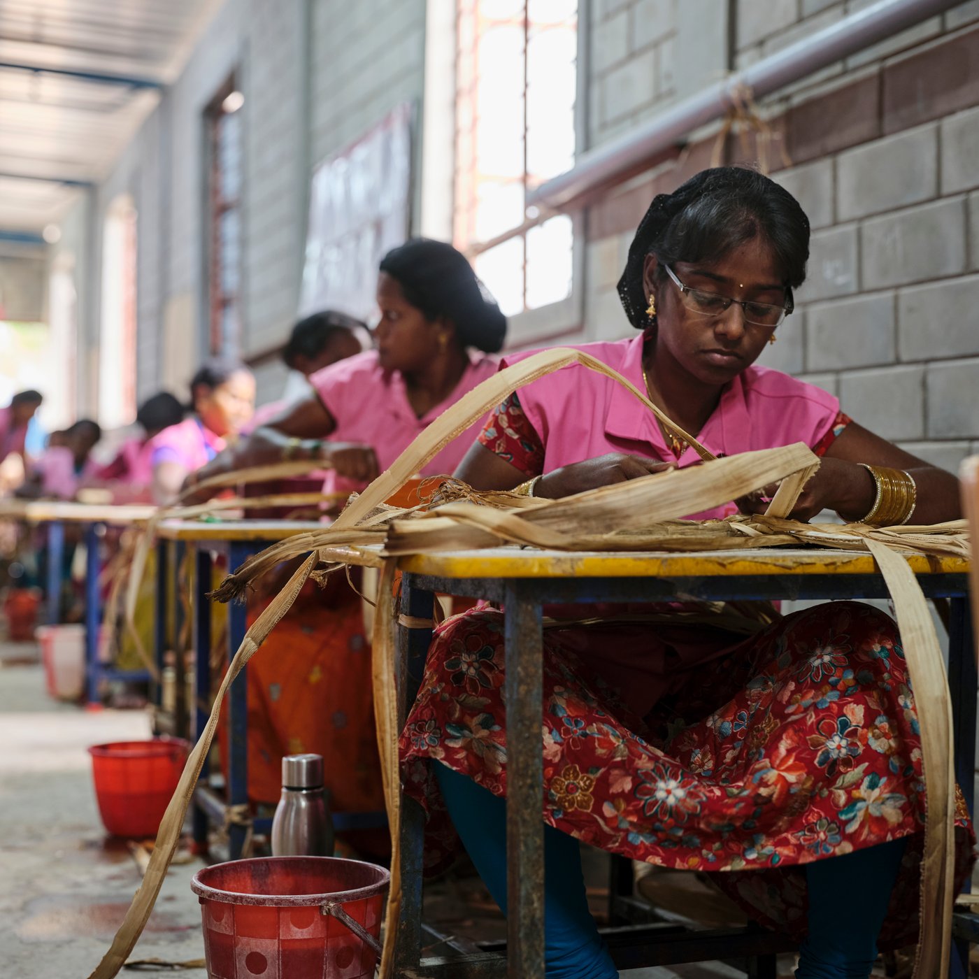 Young girls in pink clothes making baskets on small desks in a big room.