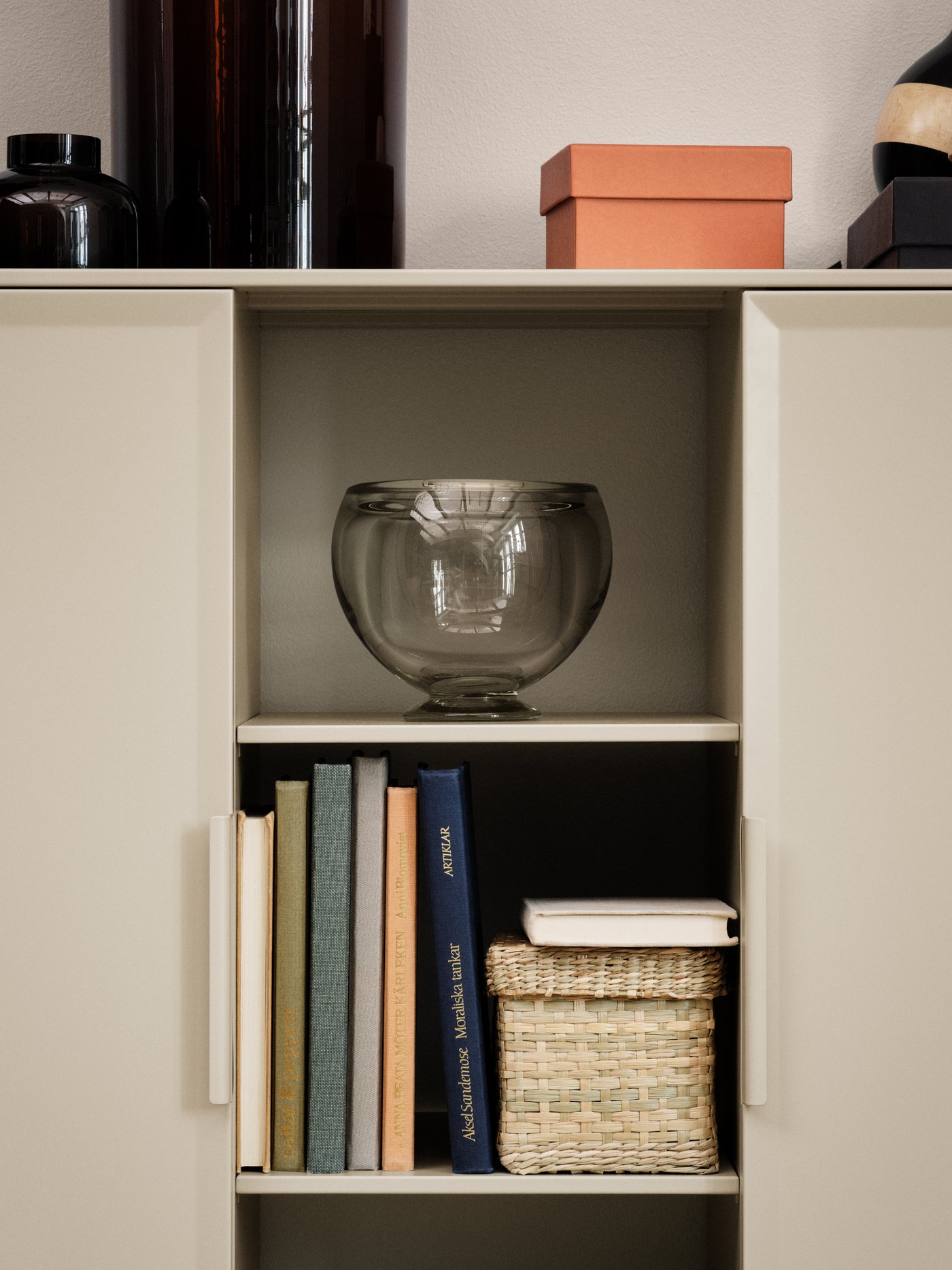 The open shelves of a beige TULLSTORP cabinet filled with a vase, some books and a basket. Boxes and glass vases are on top.