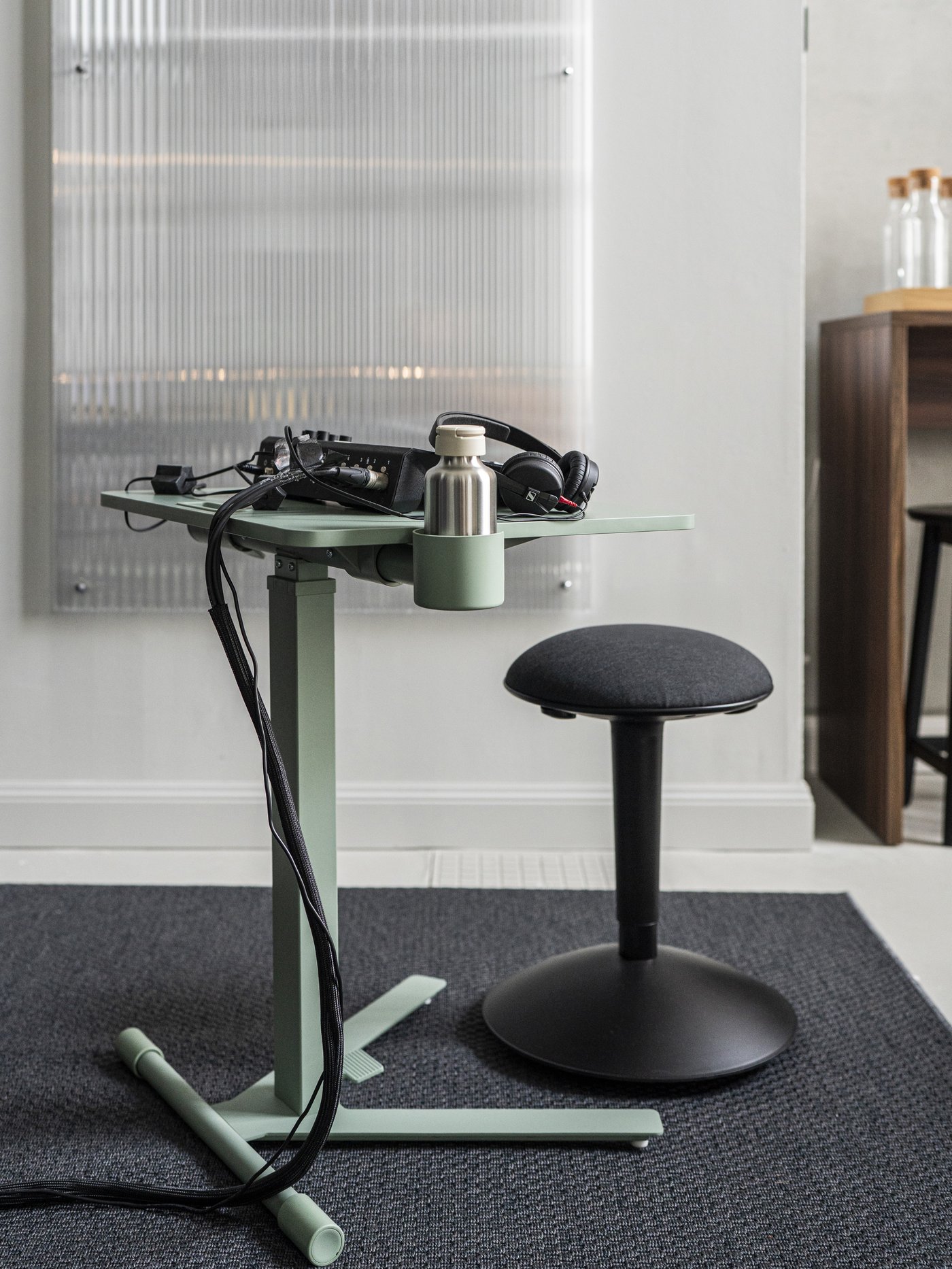 Piccolo tavolo da lavoro verde su cui sono appoggiati laptop e cavi, accanto a uno sgabello girevole nero su tappeto scuro in un ambiente moderno.